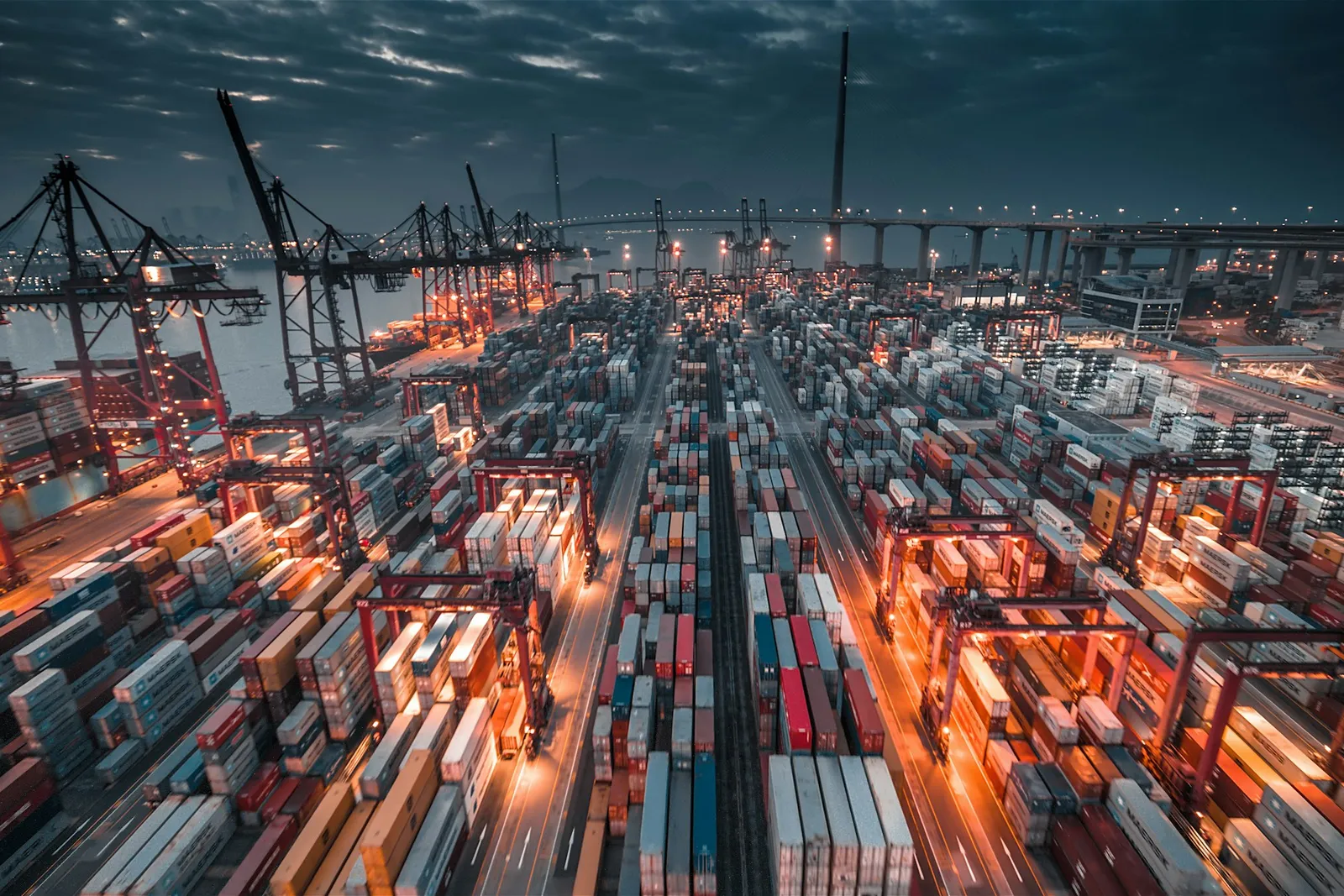 Gantry cranes and stacked shipping containers at a port under a dark sky.