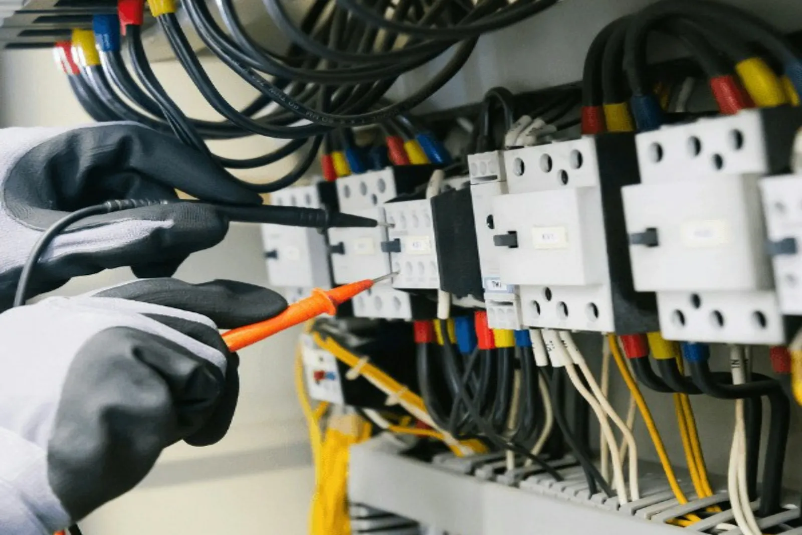 Gloved hands holding test probes against contactors inside an electrical control panel.
