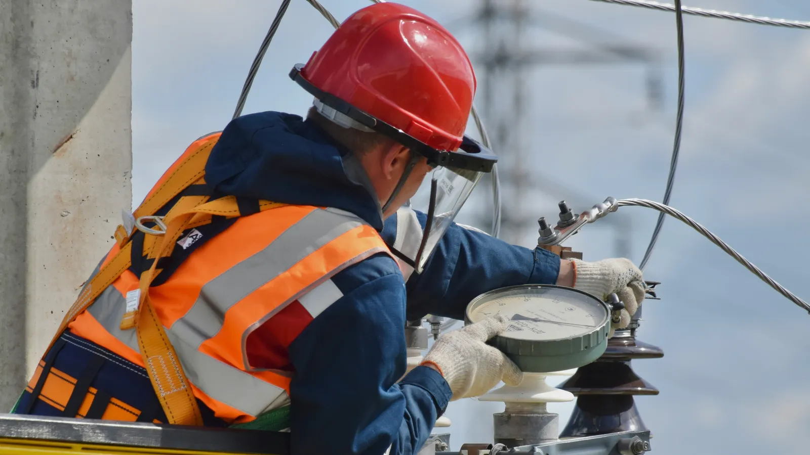 A technician in a hard hat and face shield taking a reading with a dial gauge on high-voltage equipment, power lines behind.